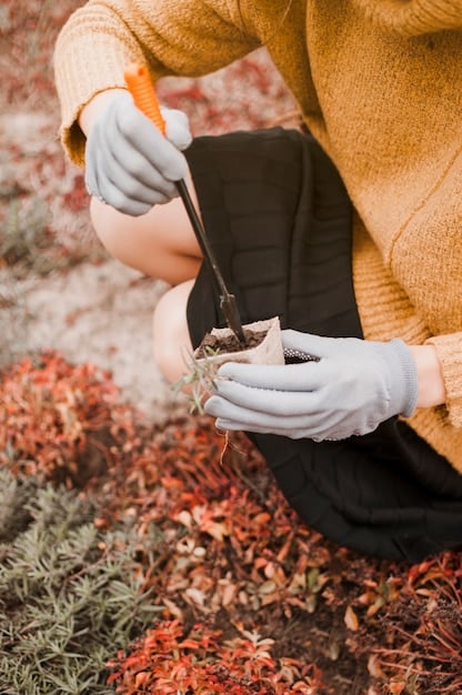 A close-up shot of someone using gardening tools to clear dry brush and leaves from around the base of a house. The homeowner is wearing gloves and protective eyewear. The image focuses on the immediate area around the house, showcasing a cleared space with exposed soil.