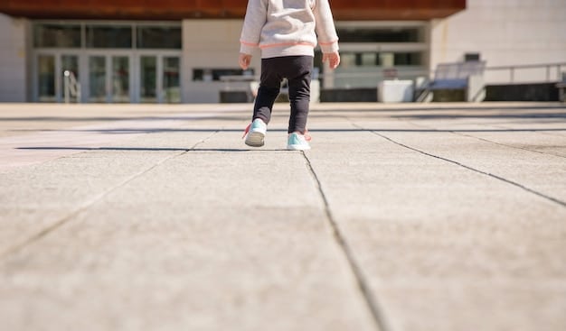 A close-up, ground-level shot of a faded crosswalk in front of a school. A pair of children's sneakers are visible stepping onto the crosswalk. Focus is on the wear and tear of the crosswalk stripes.
