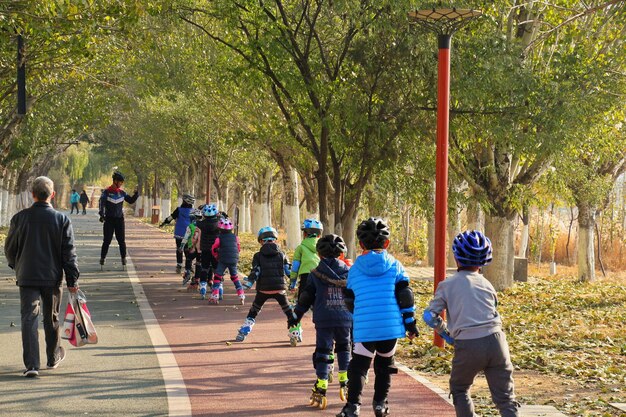 A group of children walking and biking to school, accompanied by adult volunteers. They are wearing brightly colored safety vests and helmets. The background shows a tree-lined street with minimal car traffic.