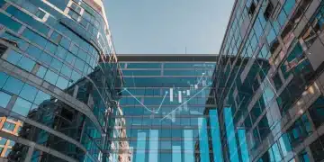 Fachada de edificio moderno en ciudad española, reflejando el cielo, con gráficos financieros ascendentes. Inversión inmobiliaria España.